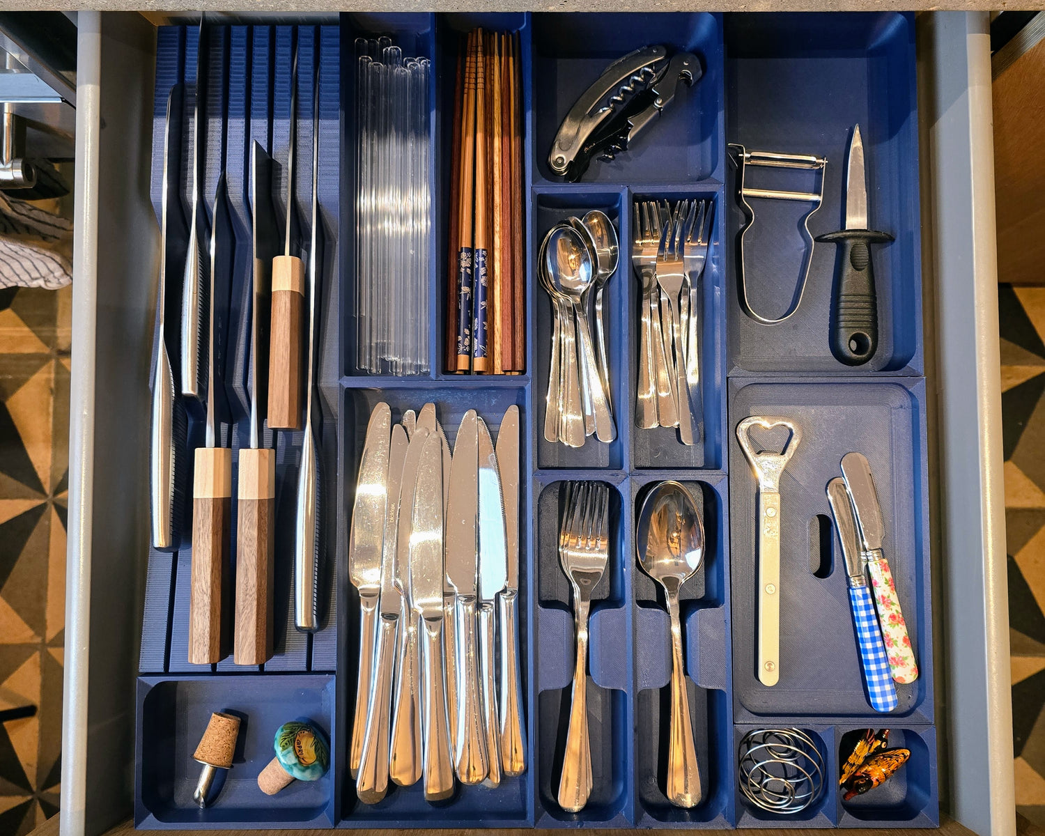 An organized kitchen drawer featuring a custom modular organizer in army blue, with neatly arranged compartments. Items include knives, forks, spoons, chopsticks, clear straws, a corkscrew, a bottle opener, a vegetable peeler, a paring knife, butter knives, and small accessories like cork stoppers and coiled bands. The organizer provides a clean, efficient layout for easy access and use.