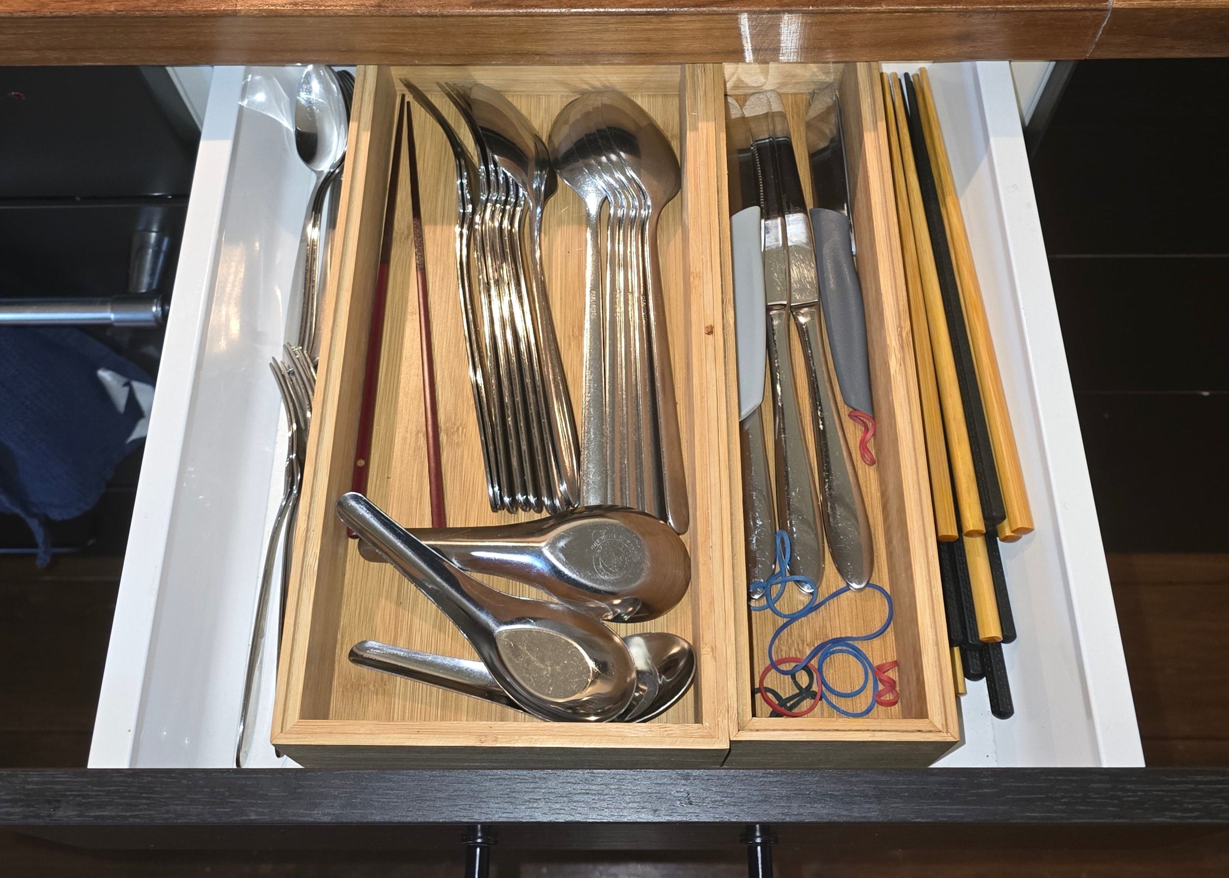 An unorganized kitchen drawer filled with wooden dividers holding cutlery and utensils. Items include spoons, forks, knives, chopsticks, rubber bands, and other kitchen tools in no specific order, showcasing a cluttered arrangement before organization.